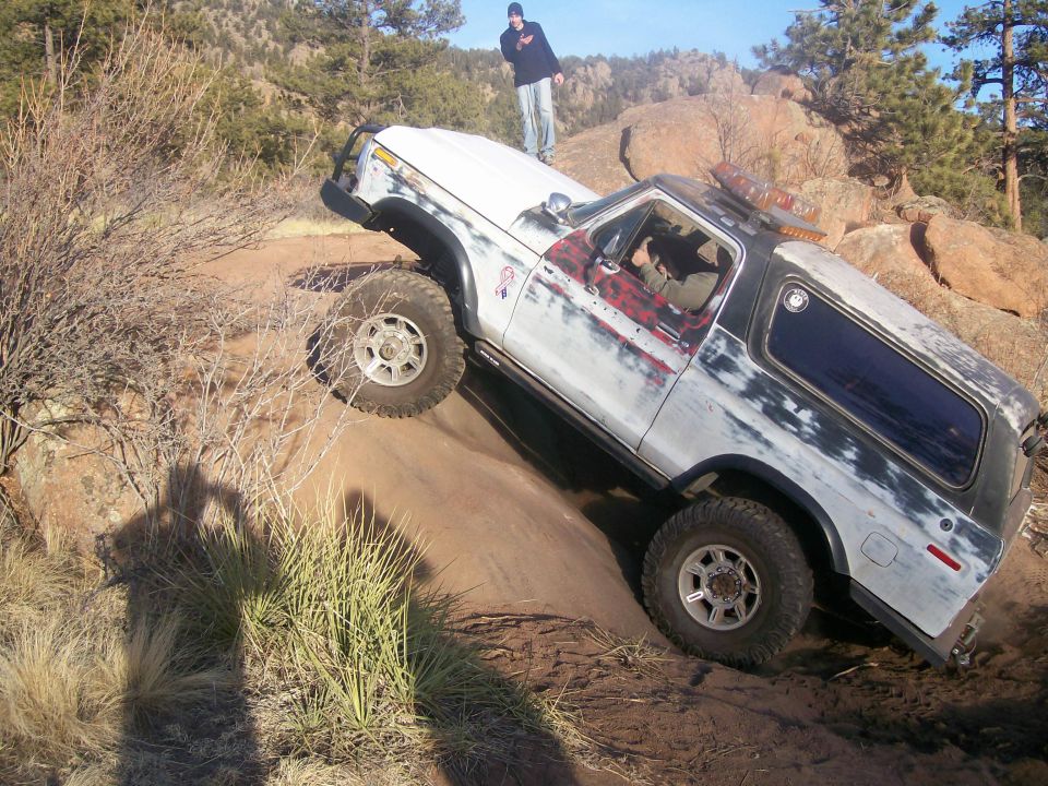 White SUV with red plaid door panel climbing a steep dirt incline, person standing on hood