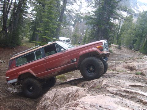 1989 Ford Bronco - This red Jeep Cherokee Chief is tackling a challenging rocky trail in a mountainous, wooded area.