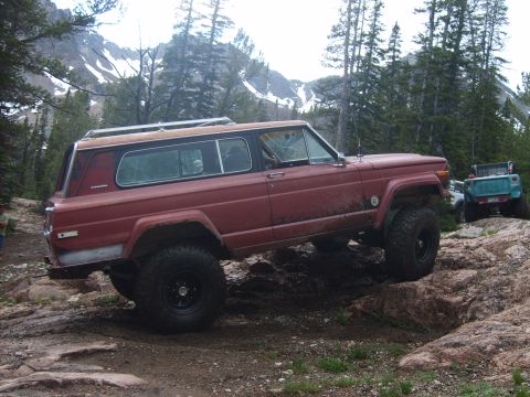 1989 Ford Bronco - A red Jeep Cherokee SJ navigates a rocky trail with snow-capped mountains in the background.