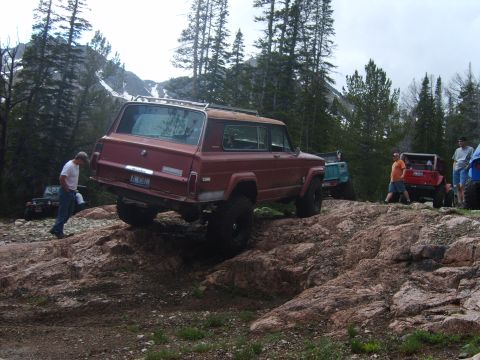 1989 Ford Bronco - Several off-road vehicles are seen on a rocky mountain trail with trees and snow-capped peaks in the background.