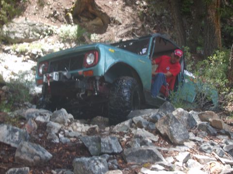1989 Ford Bronco - A man in a red shirt is driving a green off-road vehicle through a rocky, wooded area.