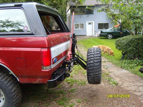 1989 Ford Bronco - Rear view of a red 1989 Ford Bronco, showing its spare tire mount and a second vehicle in the driveway.