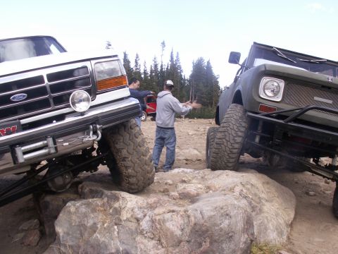1986 Ford Bronco - Two modified off-road vehicles navigate a rocky terrain with two men observing.