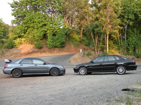 1998 Subaru Legacy - Two Subaru sedans, a gray WRX STI and a black Legacy, are parked on a gravel shoulder next to a winding road.