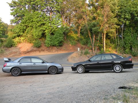 1998 Subaru Legacy - Two Subaru sedans, a gray WRX STI and a black Legacy, are parked on a gravel shoulder next to a winding road.