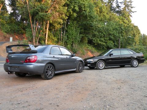 1998 Subaru Legacy - Two Subaru sedans, a gray WRX STI and a black Legacy, are parked on a gravel surface with trees in the background.