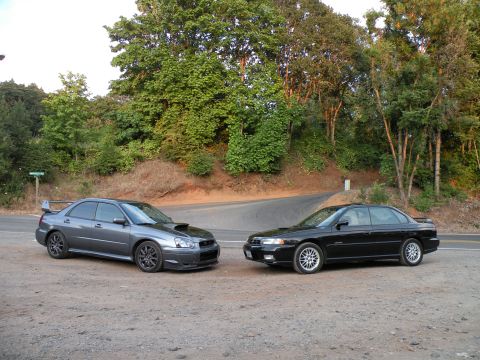 1998 Subaru Legacy - A grey Subaru WRX STI and a black Subaru Legacy are parked side-by-side on a gravel shoulder next to a winding road.