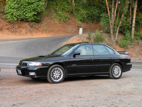 1998 Subaru Legacy - A black Subaru sedan is parked on a dirt shoulder next to a winding road.
