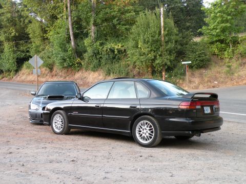 1998 Subaru Legacy - Two Subarus, a 1998 Legacy sedan and a coupe with a hood scoop, are parked on a gravel lot next to a road.