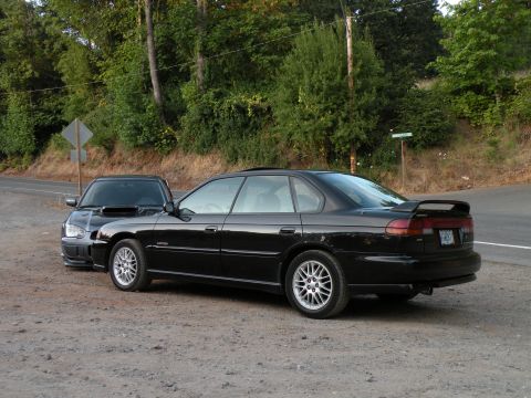 1998 Subaru Legacy - Two black Subaru sedans are parked on a gravel shoulder next to a wooded hillside.