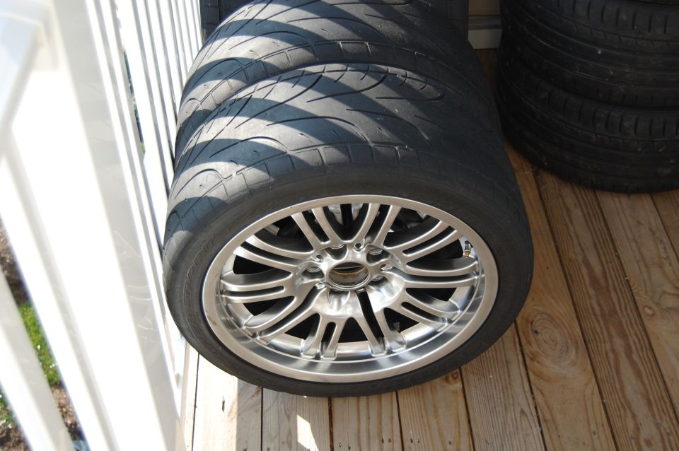 Stack of tires with silver alloy wheels. Some tires are on the ground, others are stacked on top