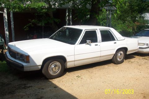 1991 Ford LTD Crown Victoria - White Ford LTD Crown Victoria four-door sedan parked on a gravel drive under shade trees, viewed from the driver's-side front three-quarter…
