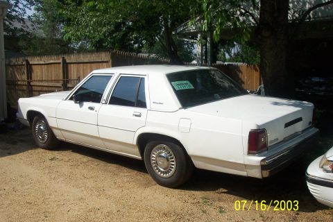 1991 Ford LTD Crown Victoria - White Ford LTD Crown Victoria four-door sedan seen from the driver's-side rear three-quarter on a gravel drive, with a green paper dealer/i…