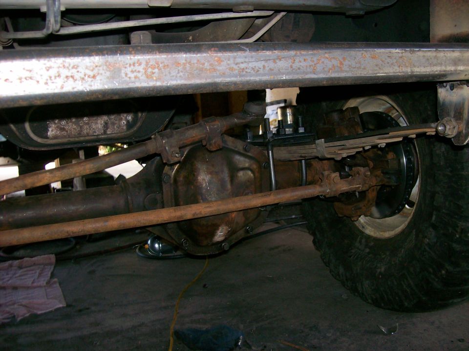 Undercarriage shot of a vehicle's front axle, steering linkage, and leaf spring suspension. A tire is visible on the right