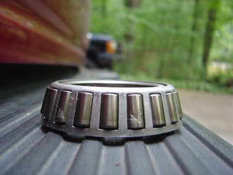 1986 Ford Bronco - A tapered roller bearing rests on a textured surface, with parts of a red vehicle and a dark SUV visible in the background.