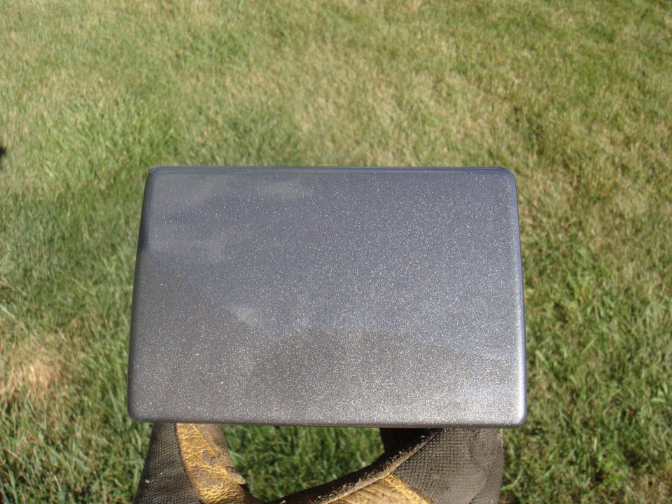 Gray metallic rectangular part held in gloved hand against green grass background
