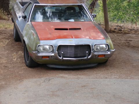 1973 Ford Ranchero - Three-quarter front view of a 1970s Ford car-based pickup in bare metal and patchy green and orange primer, parked on dirt under trees. It…
