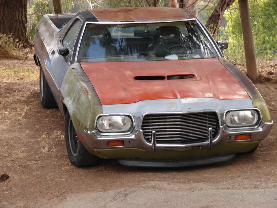 Weathered 1970s Ford Ranchero with green fenders and orange hood parked on dirt