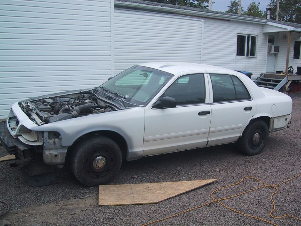 White 2003 Ford Crown Victoria with hood open, showing engine, parked on gravel in front of a white building
