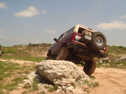 1992 Ford Bronco - A maroon and tan Ford Bronco navigates an off-road obstacle, with a spare tire and cargo carrier visible on the rear.