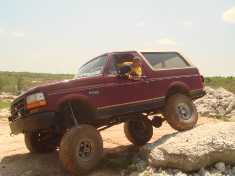 1992 Ford Bronco - A man drives a lifted maroon 1992 Ford Bronco with a tan top over rocky terrain.