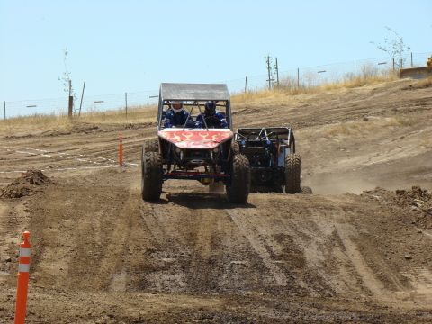 1989 Ford Bronco - Two off-road buggies compete on a dusty dirt track under a clear blue sky.