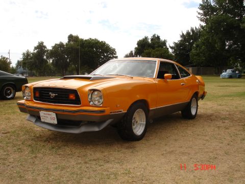 1978 Ford Mustang II - An orange coupe with a hood scoop and aftermarket wheels is parked on a grassy field.