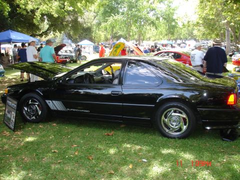 1978 Ford Mustang II - Black coupe with racing stripes and "SUPERCHARGED" text is displayed at an outdoor car show with other vehicles present.