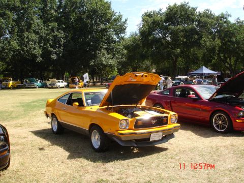 1978 Ford Mustang II - A yellow coupe with its hood open is displayed at an outdoor car show alongside other vehicles.