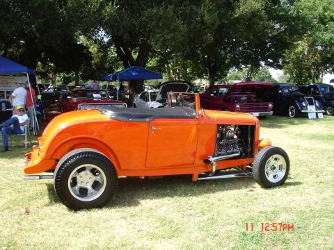 1978 Ford Mustang II - Outdoor car show features a bright orange hot rod roadster with a visible engine and other classic cars nearby.