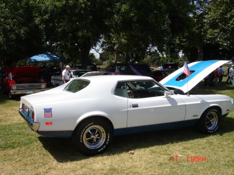 1978 Ford Mustang II - A white 1971 Ford Mustang Mach 1 with blue racing stripes and an American flag on the hood is displayed at an outdoor car show.