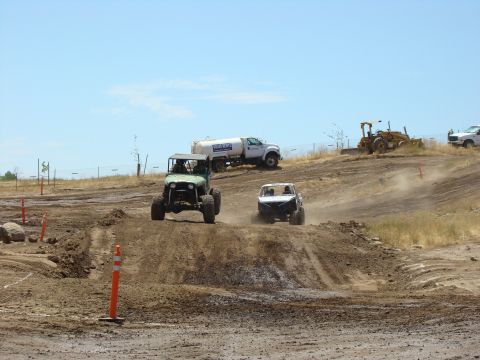 1989 Ford Bronco - Off-road vehicles compete on a dusty track under a clear blue sky, with support vehicles nearby.