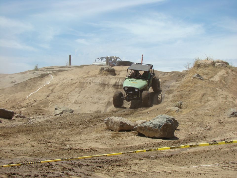 Green off-road vehicle with roll cage climbing a dirt hill, another white vehicle in background