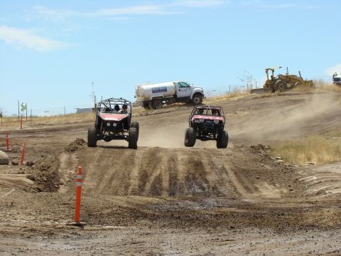 1989 Ford Bronco - Two off-road vehicles compete on a dusty track under a bright blue sky, with construction equipment visible in the distance.