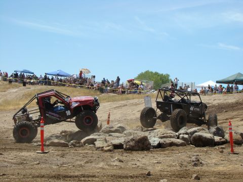 1989 Ford Bronco - Two custom off-road vehicles compete on a challenging rocky course with spectators in the background.