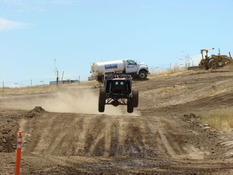 1989 Ford Bronco - An off-road buggy jumps over a dirt mound during a race, with a water truck and bulldozer visible in the background.