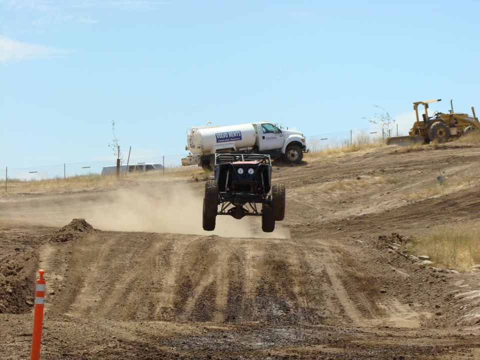 Off-road buggy airborne over dirt track, rear view. Water truck and bulldozer in background
