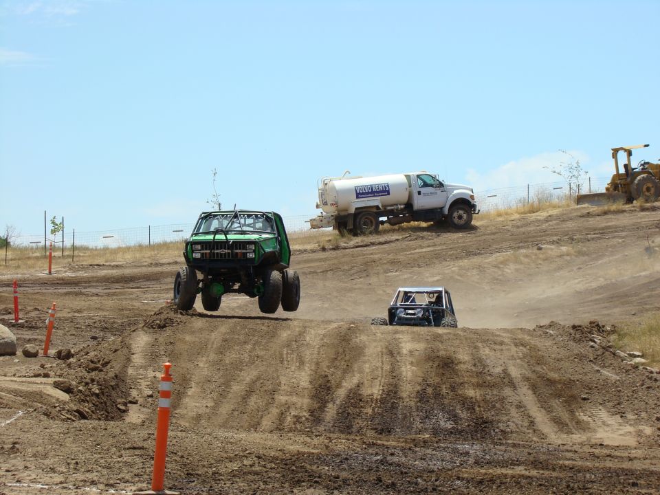 Green off-road truck airborne over a dirt mound, with a water truck and bulldozer in the background