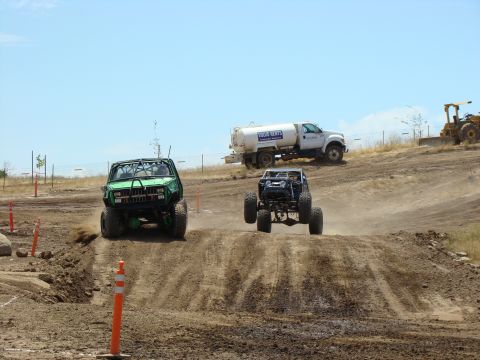 1989 Ford Bronco - Multiple off-road vehicles compete on a dusty dirt track under a clear blue sky.