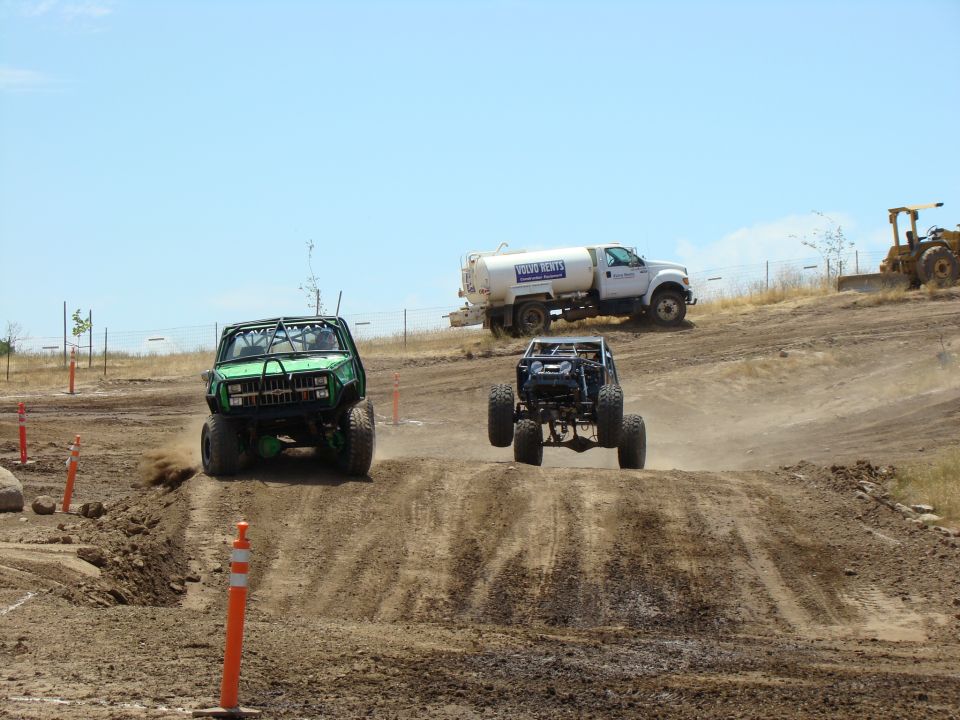 Off-road trucks race on a dirt track with a water truck and bulldozer in background