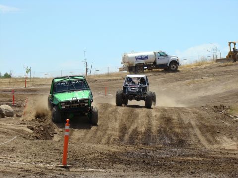 1989 Ford Bronco - Two custom off-road vehicles compete on a dusty track under a clear blue sky.