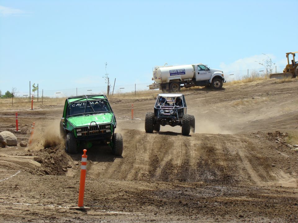 Green off-road truck with 'CAVE MAN' on top, kicking up dust on a dirt track. Another buggy follows behind. Water truck in background