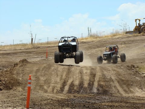 1989 Ford Bronco - Two off-road buggies compete on a dirt track, one airborne over a jump.