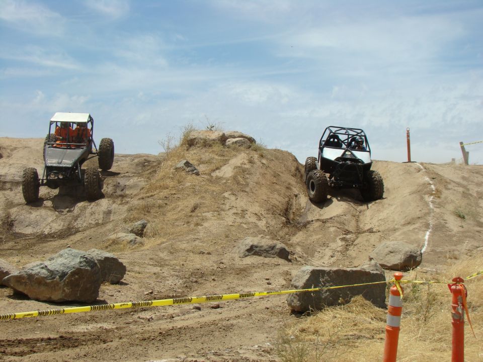 Two buggies are climbing a dirt hill with rocks and sparse vegetation under a partly cloudy sky