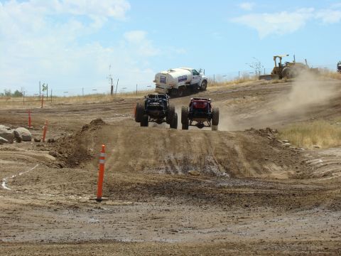 1989 Ford Bronco - Two custom off-road vehicles compete on a dusty track, kicking up dirt as they go.