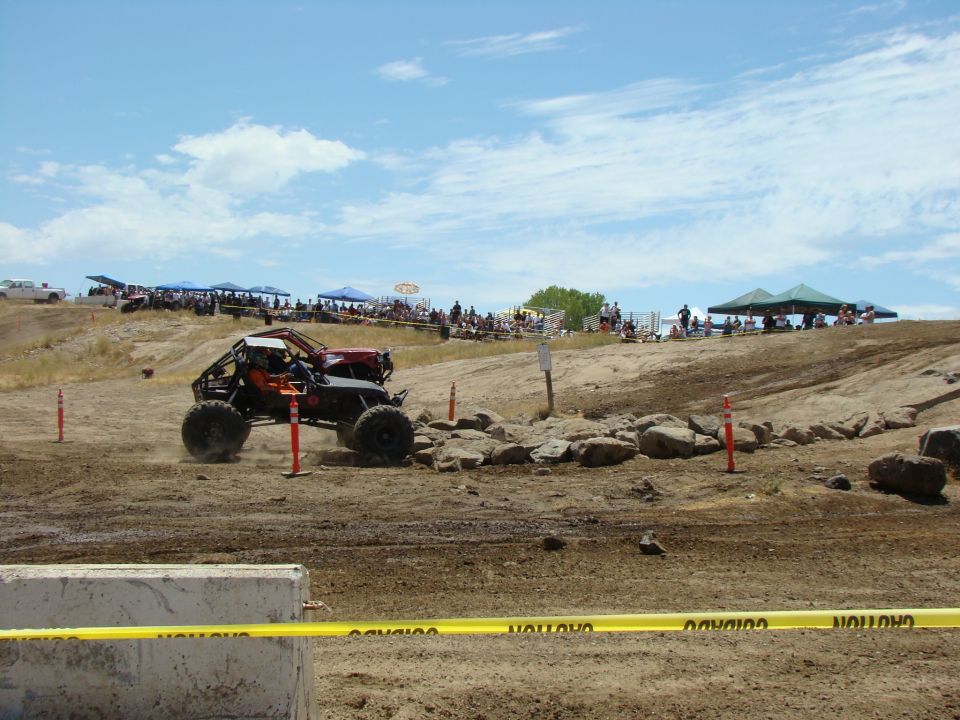 Buggy with big tires driving over rocks on a dirt course. Spectators are in the background