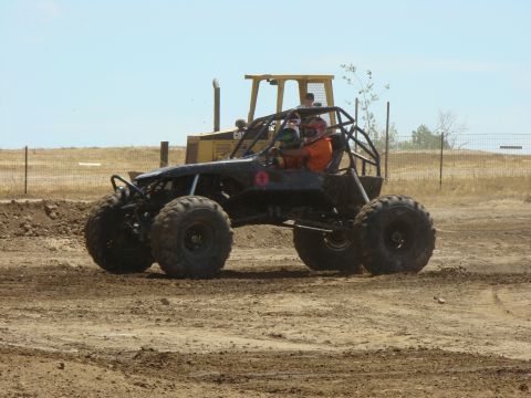 1989 Ford Bronco - Off-road buggy with roll cage and two occupants drives on dirt track, with a bulldozer behind it.