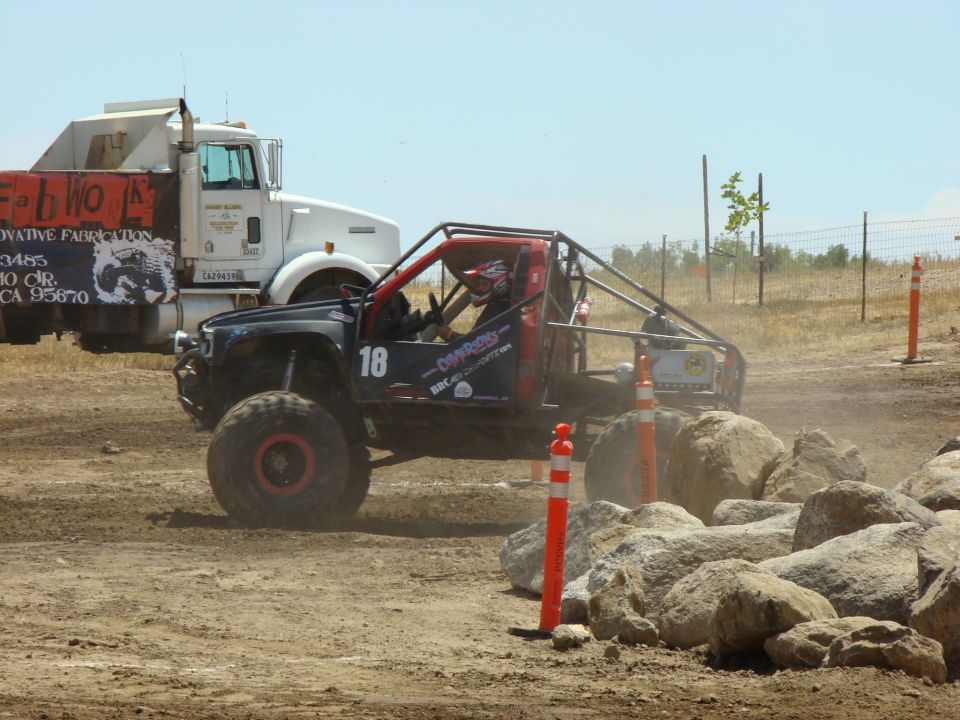 Off-road race truck with number 18 on the side navigates a rocky course. A large white truck is in the background