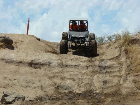 1989 Ford Bronco - Two people in an off-road buggy ascend a challenging dirt incline under a partly cloudy sky.
