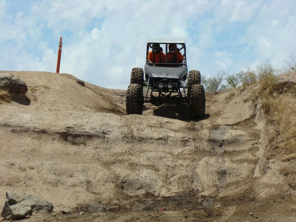 Off-road buggy with two occupants climbing a steep dirt hill. Large tires and roll cage are visible